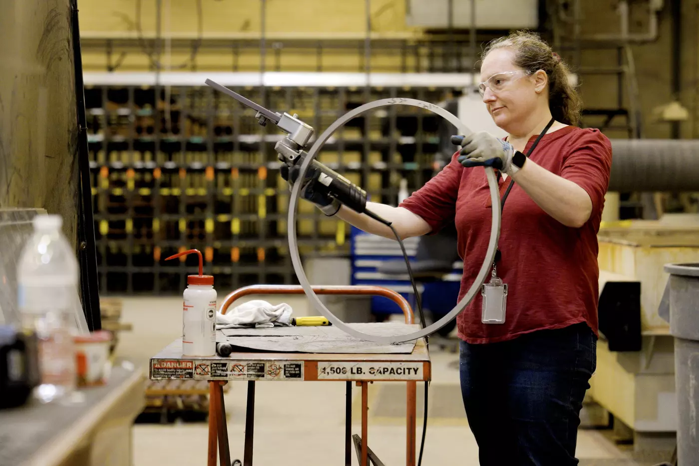 Engineering technician Kristin Lee, wearing safety glasses and gloves, uses a powered tool on a large metal ring in a machine shop