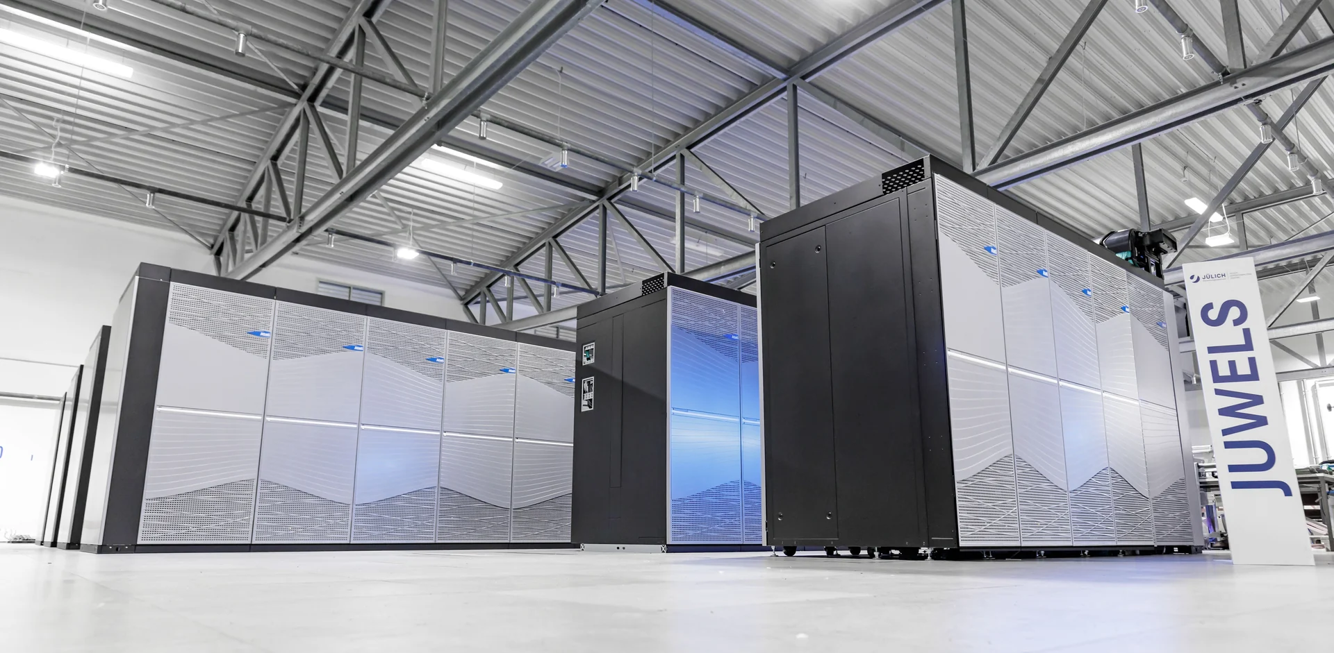 Rows of silver and black JUWELS Booster supercomputer cabinets inside a bright equipment hall.